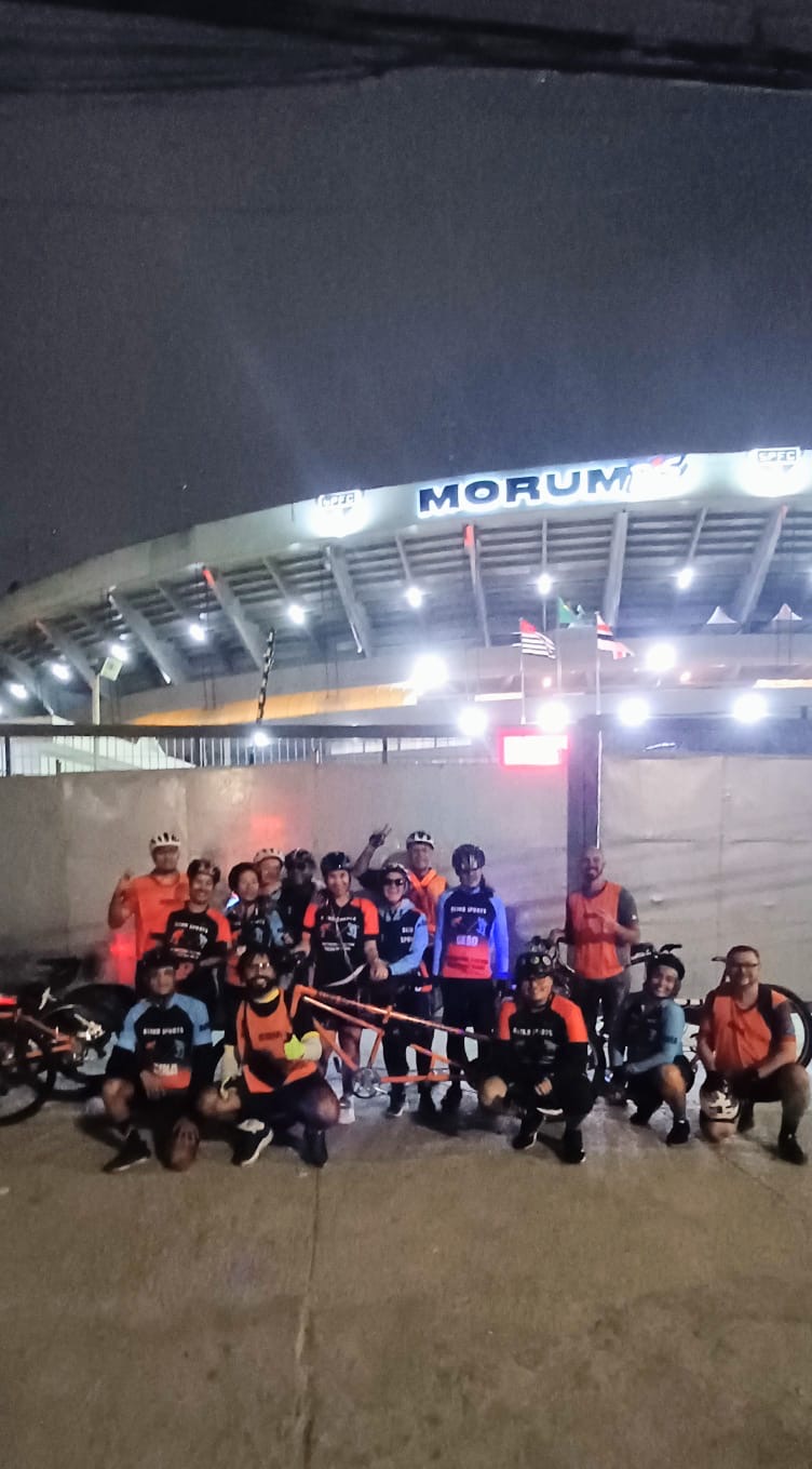 Quatro ciclistas usando roupas de ciclismo coloridas pedalando em duas bicicletas em uma rua urbana.  
À esquerda há uma bicicleta branca com duas pessoas: na frente, uma mulher de pele clara usando capacete branco, óculos escuros amarelos, camisa de manga longa azul clara com detalhes em laranja e preto, bermuda de ciclismo preta e meias azuis; ela está sorrindo e pedalando. Atrás dela está outra mulher de pele clara, também sorrindo, com capacete preto, óculos escuros, camisa azul clara com laranja e preto, bermuda preta e meias azuis.  
À direita há uma bicicleta tandem laranja, feita para duas pessoas: na frente está um homem de pele clara com barba curta, capacete preto, camisa azul clara com mangas pretas, bermuda preta e tênis azul; atrás dele está uma mulher de pele clara com cabelos escuros cacheados, capacete preto, óculos escuros, camisa azul clara, bermuda de ciclismo colorida em tons de rosa e roxo, meias verdes e tênis escuros. Entre os dois quadros da bicicleta tandem há uma bolsa cinza pendurada.  
Os quatro ciclistas parecem estar em movimento na faixa da direita de uma avenida asfaltada, com árvores e prédios ao fundo. Há também uma placa de trânsito indicando travessia de pedestres no lado direito da imagem.