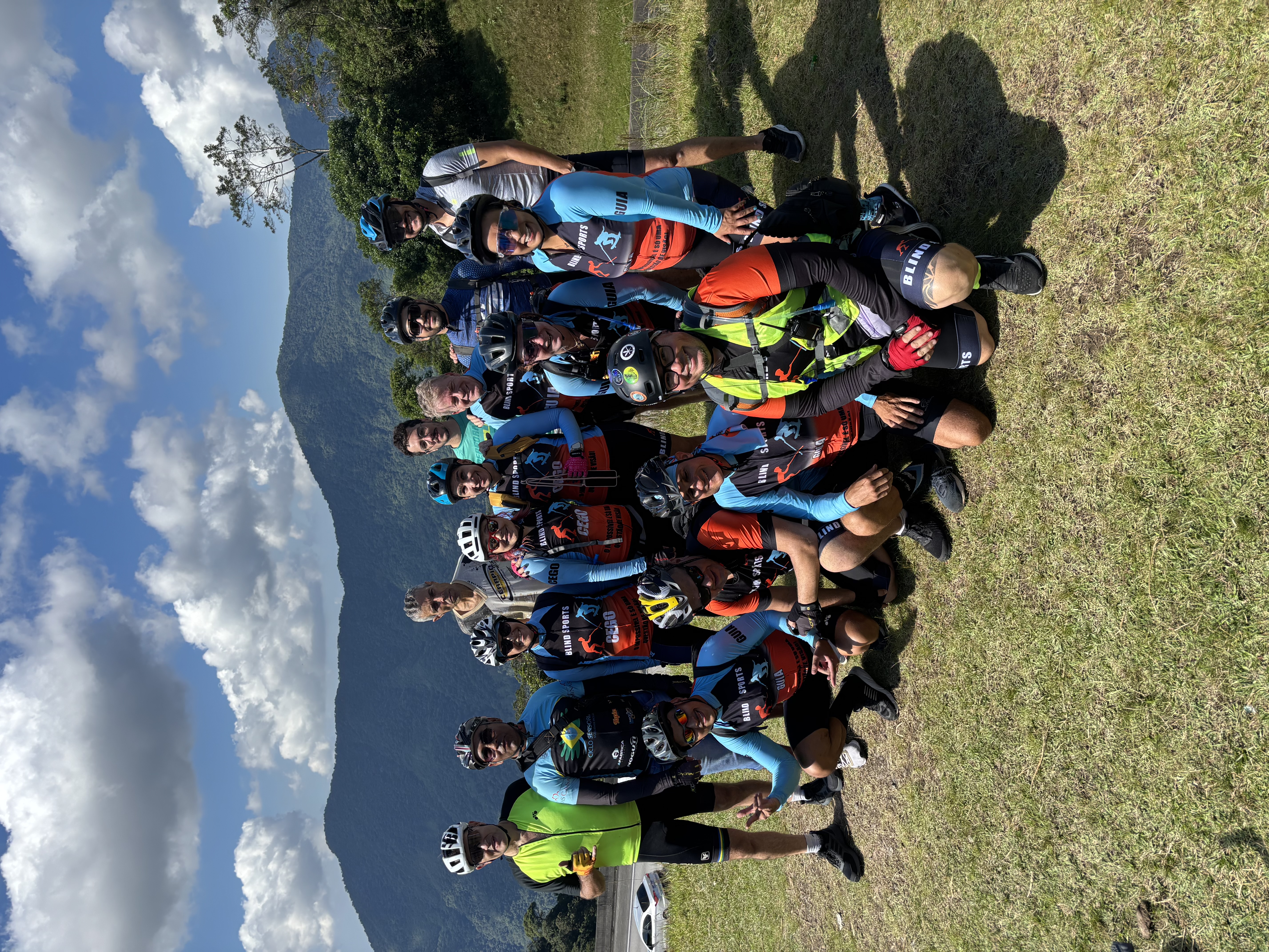 Um grupo de ciclistas posando ao ar livre em um gramado, com montanhas ao fundo sob um céu azul com nuvens brancas.  
Há cerca de 18  pessoas, a maioria usando capacetes de ciclismo e óculos escuros.  
As roupas são coloridas, predominando camisas de manga longa azul‑clara com partes em preto e laranja, e vários coletes com faixas refletivas.  
Nas camisas aparecem textos como “BLIND SPORTS” e, em alguns coletes e peitorais, as palavras “GUIA” e “CEGO”.  
Alguns ciclistas estão agachados na frente e outros em pé atrás, todos próximos uns dos outros, muitos sorrindo e fazendo gestos de alegria com as mãos, como sinal de paz e joinha.  
Ao fundo, as montanhas são cobertas de vegetação e formam uma cadeia que se estende de um lado ao outro da imagem.