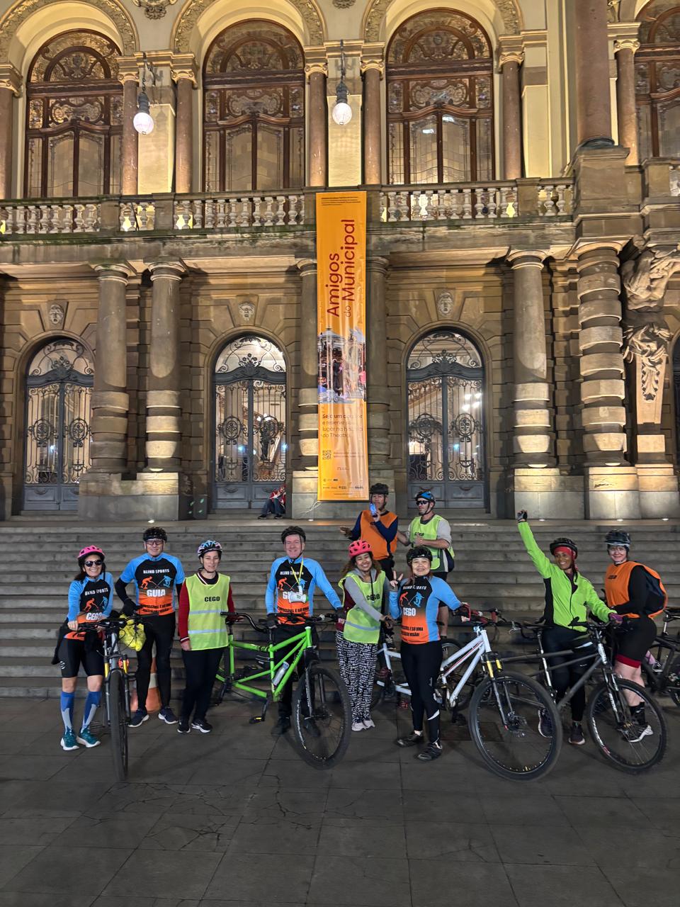 Be My AI: Grupo de pessoas com bicicletas em frente a um prédio antigo iluminado à noite. As pessoas usam capacetes e coletes refletivos, alguns azuis com texto "BLIND SPORTS CEGO" e "GUIA", outros são amarelos com a palavra "CEGO", e alguns laranja. O grupo parece estar feliz, uma pessoa levanta o braço em sinal de comemoração. Ao fundo, escadaria e portas com grades de ferro. No centro, pendurado no prédio, um banner vertical amarelo com o texto "Amigos do Municipal" em vermelho.