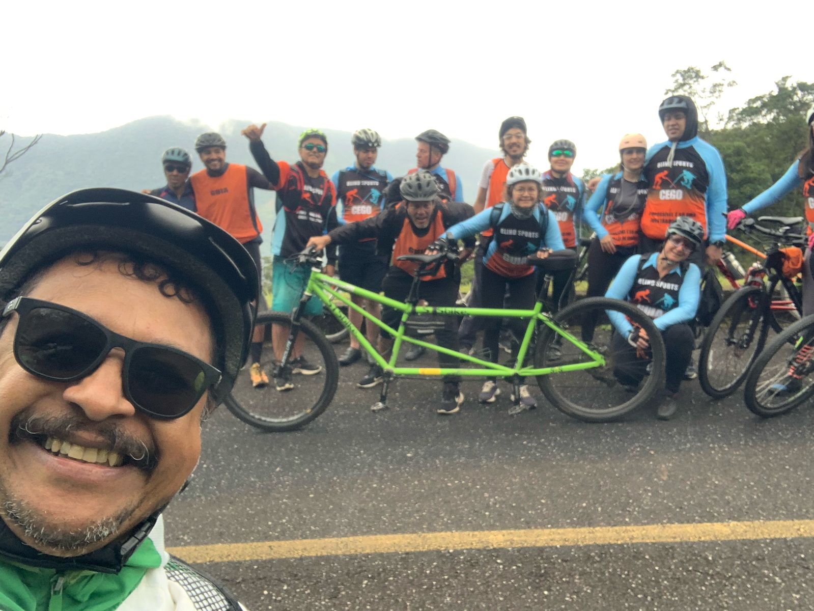 Grupo de ciclistas ao ar livre, posando para uma foto na estrada. No canto esquerdo, em primeiro plano, um homem de capacete e óculos escuros sorri para a câmera em modo selfie. Ao fundo, há aproximadamente doze pessoas, todas usando capacete e coletes de cores laranja e azul. No centro, um tandem (bicicleta dupla, verde) está sendo segurado por duas pessoas. O grupo parece animado, com alguns sorrindo e fazendo sinais positivos. Ao fundo, há vegetação e uma montanha coberta por névoa, sugerindo um ambiente natural e clima nublado.